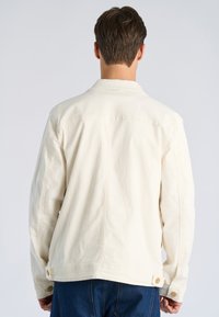 Kremowa kurtka dżinsowa o luźnym kroju, z widocznym przeszyciem, zapięciami na guziki na rękawach oraz prostym dołem. Minimalistyczny design.
