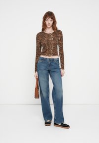 Haut à manches longues imprimé léopard avec une coupe courte, associé à un jean bleu à jambes larges. Le mannequin tient un sac à main marron et porte des chaussures noires.