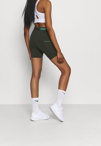 Vrouw in donkergroene, strakke fietsshorts, witte sportbeha, witte Nike-sokken en witte sneakers, zijwaarts staand tegen een effen achtergrond.