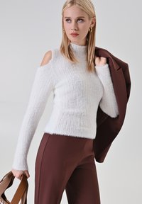 Pull en tricot blanc avec un design épaules dénudées, texture côtelée, col haut, associé à un pantalon tailleur marron et un sac à main marron.