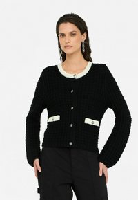 Schwarze Strickjacke mit einem weißen Kragen, großen Knöpfen und Fronttaschen. Strukturiertes Material mit einer kastigen Passform und langen Ärmeln.