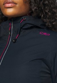 Gros plan sur une personne portant une veste bleu marine avec des cordons roses et le logo CMP sur l'épaule, montrant les lèvres et des cheveux bouclés foncés.