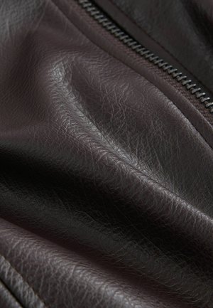 Közelkép sötétbarna bőr anyagról, amelyen látható a szemcsés textúra, és részben látható egy fém cipzár.