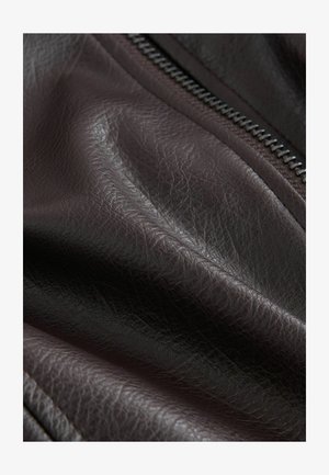 Közelkép sötétbarna bőr anyagról, amelyen látható a szemcsés textúra, és részben látható egy fém cipzár.