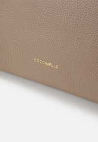 Beige nahkalaukku, jossa on teksturoitu pinta ja kultainen "Coccinelle"-logo. Minimalistinen muotoilu ilman näkyviä lisäkoristeita.