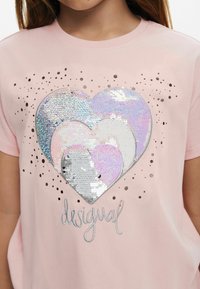 T-shirt di cotone rosa con un grande design a forma di cuore realizzato in paillettes argento, bianco e colori pastello, con accenti a pois e testo del logo.