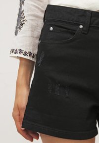 Shorts en denim noir avec un design déchiré et des détails effilochés. Comprend des clous en métal sur les poches et une légère brillance dans la texture.