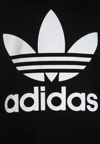 Tessuto nero con un grande logo bianco Adidas, con tre forme simili a foglie sopra e tre strisce orizzontali sotto il nome del marchio.
