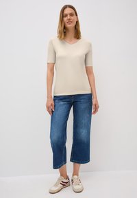 Béžová košile s krátkým rukávem a výstřihem do V v kombinaci s cropped modrými džínami a béžovými teniskami s vínovými pruhy. Jednoduchý, ležérní střih s minimálními detaily.