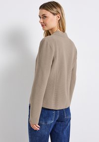 Cardigan beige con texture e design strutturato, maniche lunghe e scollo rotondo, abbinato a jeans in denim blu.