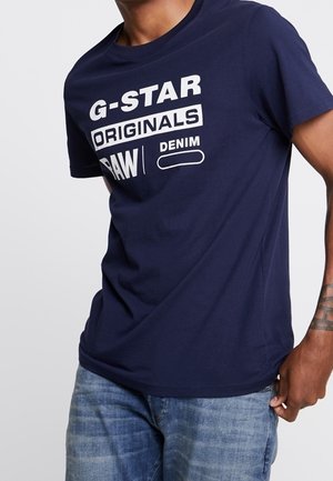 Mand iført en mørkeblå T-shirt med hvid tekst, der lyder G-Star Originals Raw Denim, og lyseblå jeans.