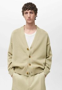 Beige stickad kappa med V-krage, långa ärmar och tre bruna knappar. Texturerad tyg med ribbade manschetter och fåll. Bärs över en vit skjorta.