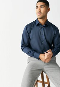 Młody mężczyzna z krótkimi ciemnymi włosami, ubrany w granatową koszulę zapinaną na guziki oraz jasnoszare spodnie, siedzi na drewnianym stołku na białym tle.