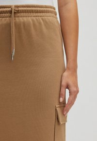 Pantalons de survêtement en mélange de coton de couleur marron clair avec une taille élastique et un cordon de serrage. Présente une poche plaquée sur le côté. Texture lisse.