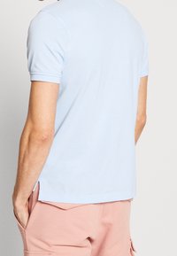 Ljusblå t-shirt med kort ärm och rund halsringning, har en mjuk textur och sidoflikar vid fållen. Bärs med rosa shorts.