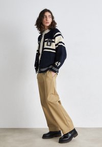 Cardigan bleu marine et crème avec motifs rayés et détails lettres, associé à un pantalon beige et des bottes noires, présentant une coupe décontractée.