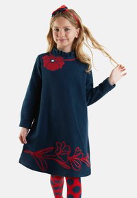 Junges Mädchen in einem marineblauen Kleid mit roten Blumendesigns, roten und blauen gepunkteten Leggings und einem roten Haarband mit einer Schleife.