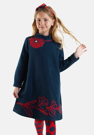 Meisje in een marineblauwe jurk met rode bloemenpatronen, rode en blauwe stippenleggingen, en een rode haarband met een strik.