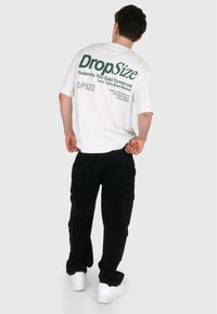 T-shirt blanc à manches courtes avec du texte vert au dos, associé à un pantalon cargo noir. Le modèle porte des baskets blanches.