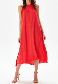 Robe maxi rouge sans manches avec un col haut et un tissu texturé ; présente une silhouette fluide et un ourlet légèrement plus court à l'avant.