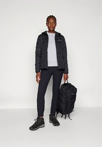 Sort dunjakke med hætte, grå sweater, sorte bukser og sorte vandrestøvler. Holder en sort rygsæk med mærke.