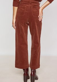 Pantaloni larghi in corduroy ruggine con vita alta, dotati di tasche posteriori e superficie testurizzata, abbinati a stivaletti bordeaux con tacco.