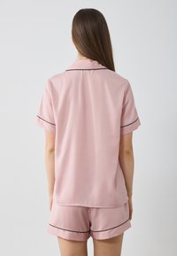 Korte roze pyjama-set met een geknoopte korte mouw shirt en shorts, met zwarte piping langs de randen voor contrast. Eenvoudige textuur.