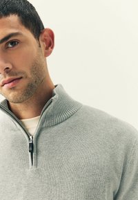 Primer plano de un hombre con cabello corto y oscuro, vistiendo un suéter gris claro con cremallera sobre una camisa blanca, mirando hacia adelante contra un fondo liso.