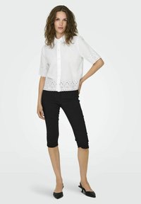 Blouse blanche à manches courtes avec boutonnage sur le devant et détail en œillets à l'ourlet, associée à un legging capri noir et des ballerines noires à bout pointu.