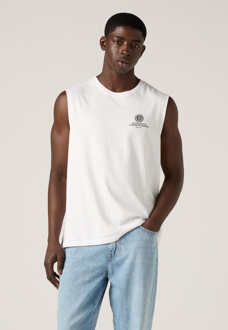 Jonge man met een wit mouwloos shirt met een klein logo en lichtblauwe jeans, staand met één hand in de zak.