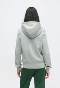 Persona ar gariem tumšiem matiem, ģērbusies gaiši pelēkā kapučjakā un tumši zaļās biksēs, stāv ar muguru pret vienkāršu baltu fonu.