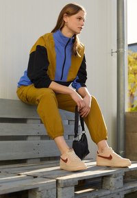 Colorful windbreaker in yellow, blue, and black; relaxed-fit tan pants; cream sneakers with red accents; sitting on a wooden bench.