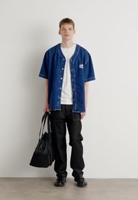 Denim kortermet skjorte, blå, med knapper og logo; hvit t-skjorte under; svarte bukser; svarte sko; bærer en svart veske.