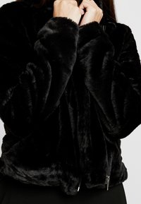 Person wearing a plush black faux fur jacket with a front zipper, holding the collar close to their neck.