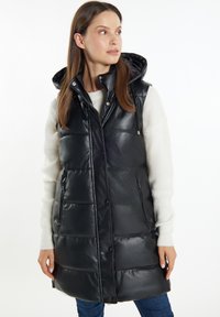 Sort polstret vest med glansfuld overflade, vedhæftet hætte og snaplukning foran. Har to lynlåslommer på siden. Bæres over en cremet sweater.
