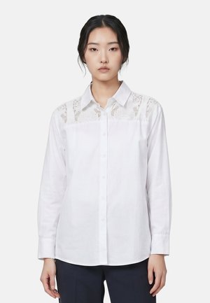 Femme aux cheveux noirs portant une chemise blanche à boutons avec des détails en dentelle sur les épaules et un pantalon sombre, debout devant un fond uni.