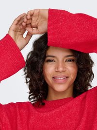 Pull rouge à coupe décontractée et manches larges. Tricot doux et texturé avec un léger éclat, porté avec les bras levés, révélant une peau lisse.