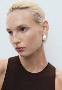 Boucle d'oreille argentée en forme abstraite, surface lisse et brillante. Modèle avec des cheveux blonds clairs et une peau claire, portant un haut sans manches marron.