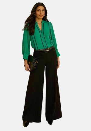 Žena v zelenej blúzke s čiernymi zvislými pruhmi, čiernych širokých nohaviciach, čiernom opasku, čiernej kabelke a čiernych špicatých topánkach, stojaca.