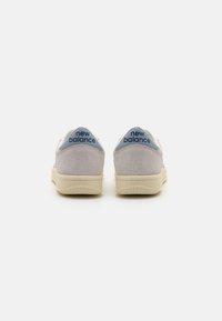 Adidași din suede gri cu o talpă crem, având un tab de călcâi de culoare albastră deschisă inscripționat cu "new balance". Designul este low-top și minimalist.