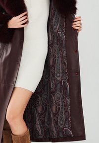 Cappotto in pelle bordeaux con colletto in pelliccia sintetica, dotato di una fodera a motivo paisley in profondo viola e rosso, che presenta eleganti dettagli con bottoni.
