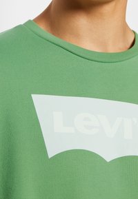 Gros plan sur le cou et le haut de la poitrine portant un t-shirt vert à col rond avec un logo Levi's blanc partiel.