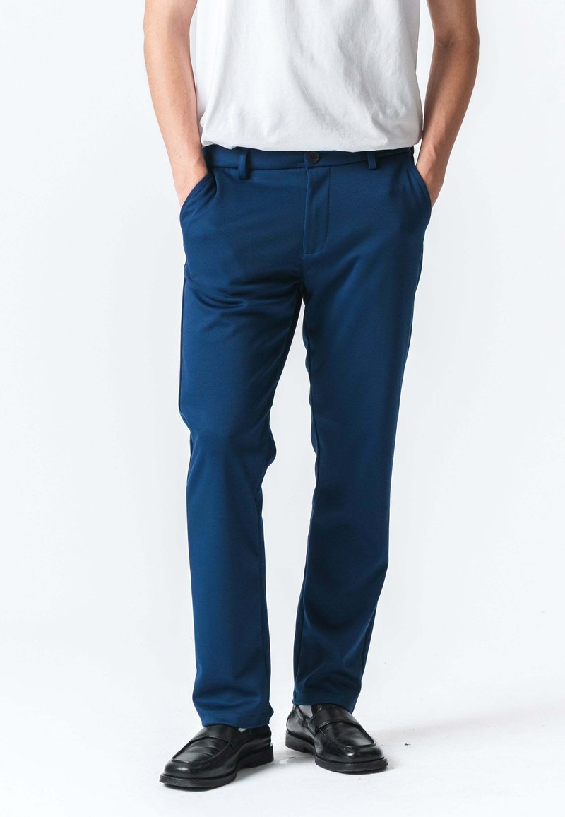 Marineblå bukser med en glat tekstur, slim fit design og forlommer. Båret med en ensfarvet hvid t-shirt og sorte sko.