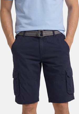 Mann trägt marineblaue Cargoshorts und einen grauen Stoffgürtel, mit den Händen in den Seitentaschen, vor einem schlichten Hintergrund.