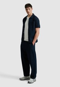 Marineblaues, strukturiertes Kurzarmhemd mit einer Brusttasche, über einem weißen gerippten Strickshirt getragen. Locker sitzende dunkle Hose und graue Sneakers.