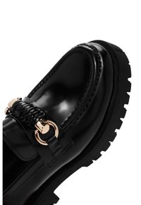 Mocassino in pelle nera con un accento intrecciato e hardware color oro. Presenta una suola in gomma spessa con battistrada testurizzato per una migliore aderenza.