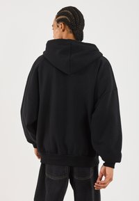 Hanorac negru cu glugă mare, croială lejeră, umeri lăsați și manșete cu risc. Textură a materialului netedă și tiv elastic. Poartă cu blugi închis la culoare.