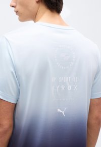 Άνδρας που φορά μπλούζα αθλητική Puma με διαβάθμιση από ανοιχτό μπλε σε σκούρο μπλε, με το κείμενο «My Sport is HYROX» και «fitness racing series» στην πλάτη.