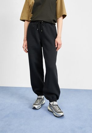 Person wearing black drawstring sweatpants with elastic cuffs and silver-white sneakers standing on a light blue floor against a white wall.