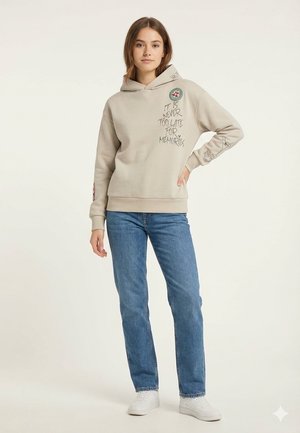 Junge Frau steht mit der Hand in der Hüfte, trägt einen beigen Hoodie mit dem Schriftzug "Es ist nie zu spät für Erinnerungen" und blaue Jeans mit weißen Turnschuhen.